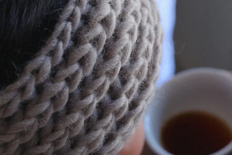 Žena pije výběrovou kávu a má na sobě pletenou čelenku. Pletená Čelenka vzorem Brioche z přírodní příze Snow od Norské značky DROPS upletená díky DIY Pletací Box