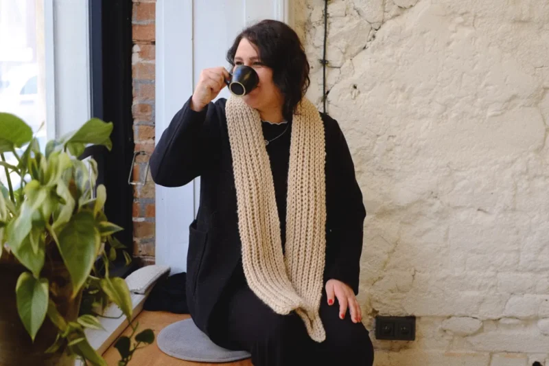 Slečna sedí v kavárně a pije kávu, na sobě má bílou pletenou šálu z příze Andes od značky DROPS.