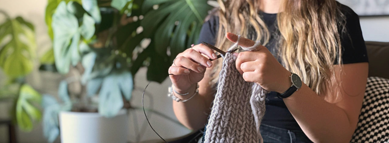 žena sedí v křesle a plete pletenou čelenku, v ruce drží kruhové jehlice a přízi Snow od Norské značky DROPS. V pozadí je list pokojové rostliny Monstera Deliciosa